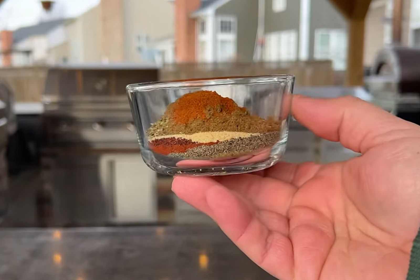 A small glass bowl of layered BBQ rub spices held in an open hand in front of an outdoor grill kitchen